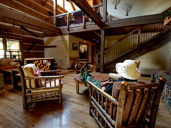 Great room looking up at the balcony, classic wooden fruniture and tables round the central fireplace with a chimney goint up throught the a-frame ceiling