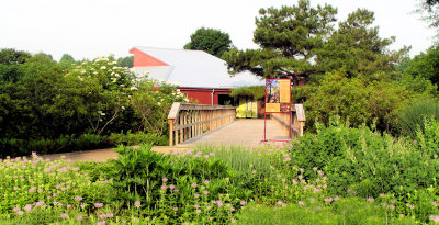 Wooden bridge walkway into the red colored arboretum building in the summer with lots of green plants and flowers.