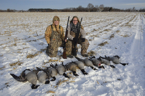 Two women in the snow holding guns with a row of dead geese lying in front of them.